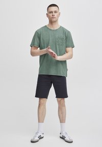 Camiseta de algodón verde con un bolsillo en el pecho, combinada con pantalones cortos oscuros y calcetines blancos. El modelo está de pie con las manos entrelazadas, frente a un fondo liso.