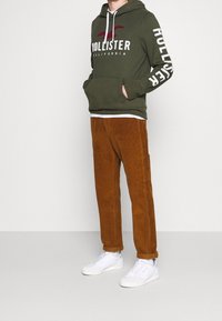 Person klädd i en grön Hollister-hoodie med vita snoddar, bruna kordflamsbyxor och vita sneakers, stående mot en neutral bakgrund.