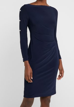 Vrouw draagt een nauwsluitende marineblauwe jurk tot op de knie met lange mouwen en decoratieve knopen langs de buitenkant van één mouw.