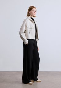 Κοντή μπεζ ζακέτα με εμπρόσθια τσέπη, σε συνδυασμό με μαύρο παντελόνι φαρδύ. Το μοντέλο στέκεται σε λείο γκρι δάπεδο, φορώντας ανοιχτά αθλητικά παπούτσια.