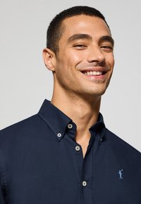 Mörkblå skjorta med knappar i en mjuk textur, med krage och ljusblå logobroderi. Knapparna är grå med en slät yta.