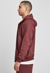 Burgundy vetoketjullinen huppari, jossa on nyörillinen huppu, kengurutasku, ribbaus hihansuissa ja helman alareunassa, valmistettu pehmeästä puuvillamateriaalista.