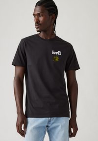 Sort bomulds-t-shirt med en hvid Levi's-logo og et lille grønt grafisk motiv på brystet. Korte ærmer og rund halsudskæring.