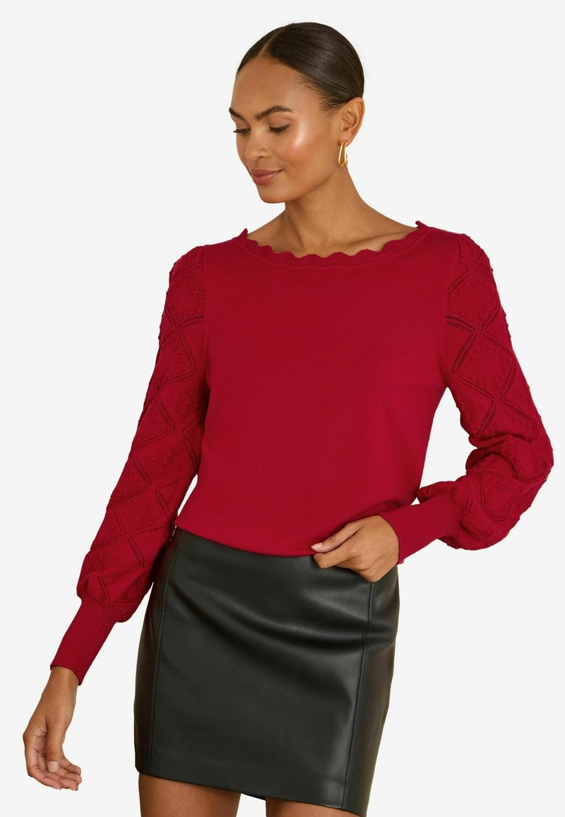Vrouw met een rood longsleeve top met getextureerde mouwen en een gegolfde halslijn, gecombineerd met een zwarte leren minirok.