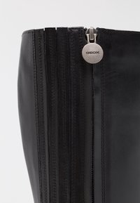 Botte en cuir noir haute jusqu'au genou avec des accents de couture verticale et une fermeture éclair argentée arborant un logo GEox rond. Texture lisse et design élégant.