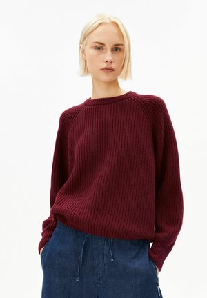 Junge Frau mit kurzem blondem Haar, die einen strukturierten bordeauxroten Pullover und blaue Jeans trägt, steht vor einem schlichten weißen Hintergrund.