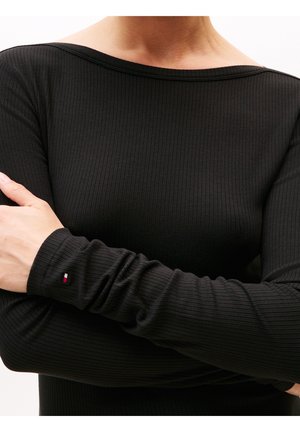 Person trägt ein enganliegendes schwarzes, geripptes Langarmshirt mit einem kleinen rot-weiß-blauen Logo am linken Handgelenk, Arme verschränkt.