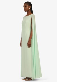 Robe maxi vert menthe avec des manches en cape, dotée d'un décolleté bateau et de poches latérales. Texture lisse, design longueur sol, associée à des chaussures ornées.