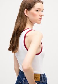 Débardeur blanc avec des bordures bordeaux, design ajusté, texture côtelée, porté avec un jean bleu taille haute avec un patch logo visible.