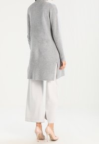 Lång grå stickad cardigan med sidoklumpar, bärs över vita vida byxor och nakna klackar. Mjuk textur och avslappnad passform.