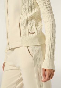 Cardigan in maglia cable-knit color avorio con polsini e orlo a coste, dotato di chiusura a zip e una piccola etichetta marrone con "FREDDY" in vita.