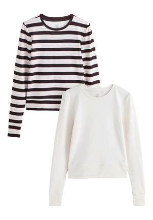 Fiatal nő hosszú barna hajjal, fekete-fehér csíkos hosszú ujjú pólót és világoskék farmert visel, kezek a zsebekben, egyszínű háttér előtt.