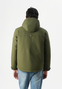 Olivegrüner Kapuzenjacke mit glatter Textur, minimaler Markierung auf der Rückseite, kombiniert mit hellblauen Jeans.