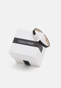 Black leather bracelet with silver clasp on a white box featuring a black stripe and "Calvin Klein" logo. Smooth texture, minimalist design.