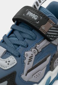 Baskets bleues avec un matériau synthétique texturé, une sangle noire contrastante avec un label "PMG" et des accents imprimés détaillés en gris et marron.