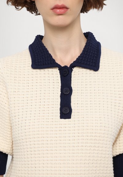 Polo de couleur crème avec une texture, un col bleu marine et une patte de boutonnage à trois boutons. Le matériau présente un motif côtelé et des manches courtes.