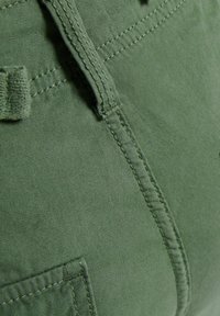 Zielony materiał z wyraźnymi szwami, pętlą na pasek i częścią kieszeni; gładka tekstura z lekko matowym wykończeniem.