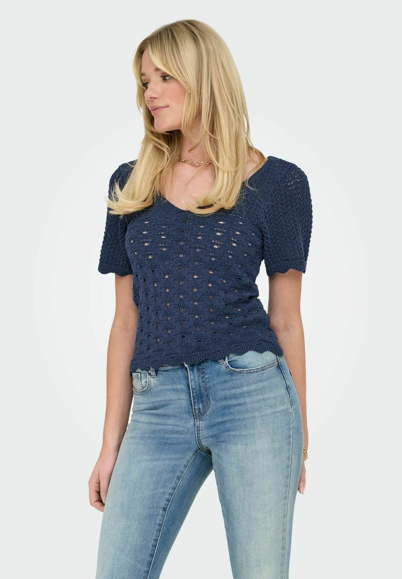 Jūras zilas krāsas adīts tops ar īsām pūkainām piedurknēm, kas ir ar atvērtu aušanu un viļņotu apakšmalu. Valkāts ar gaiši ziliem džinsiem.