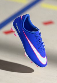 Μπλε ποδοσφαιρικά παπούτσια Nike Mercurial με άσπρες και ροζ λεπτομέρειες, να αιωρούνται πάνω από ένα ανοιχτόχρωμο πάτωμα με μπλε και κόκκινες γραμμές.