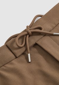 Pantalon en tissu marron avec un cordon de serrage noué avec des embouts métalliques à la taille, sur un fond clair uni.