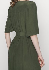 Olive green dress with short sleeves, a textured fabric, and a belted waist. Features a gathered shoulder design and flowing silhouette.