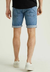 Šviesiai mėlyni džinsiniai šortai, su sukritusiais kraštais, užpakaliniai kišeniai su įstrižais siūlais, dėvimi su baltais sportbačiais neutraliame fone.