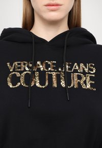 Zwarte hoodie met een gouden, met pailletten versierde logo dat "VERSACE JEANS COUTURE" leest, met een capuchon met trekkoord. Soepele stofstructuur.