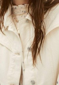 Weiße Jeansjacke mit silbernen Knöpfen, die einen strukturierten Kragen aufweist. Darunter ist eine leicht gemusterte Bluse mit fransierten Kanten geschichtet.