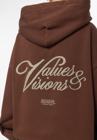 Sudadera marrón con un gran texto gráfico en crema "Valores y Visiones" en la parte trasera, con un logotipo más pequeño debajo en un color similar.
