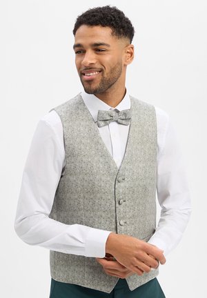 Lächelnder Mann, der eine hellgraue gemusterte Weste und eine passende Fliege über einem weißen Hemd trägt und seine Hände vor sich anpasst.