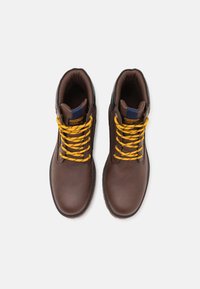 Botas de tobillo de cuero marrón con cordones amarillos y negros con un patrón, cuello acolchado y una etiqueta azul en la lengüeta. Suela de goma con agarre.