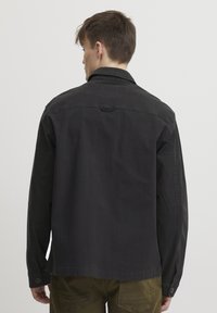 Black cotton jacket with long sleeves, a point collar, and buttoned cuffs, featuring a relaxed fit and subtle texture details on the fabric.