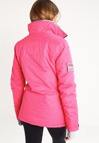 Veste isolante rose avec un col haut et un intérieur gris, arborant un patch logo sur la manche gauche et un design ajusté.