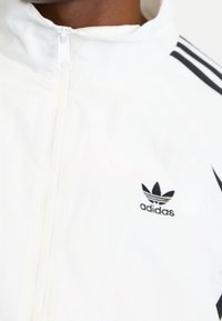 Vit zip-up jacka i lätt material, med svart Adidas-logga och tre svarta ränder på ärmarna.