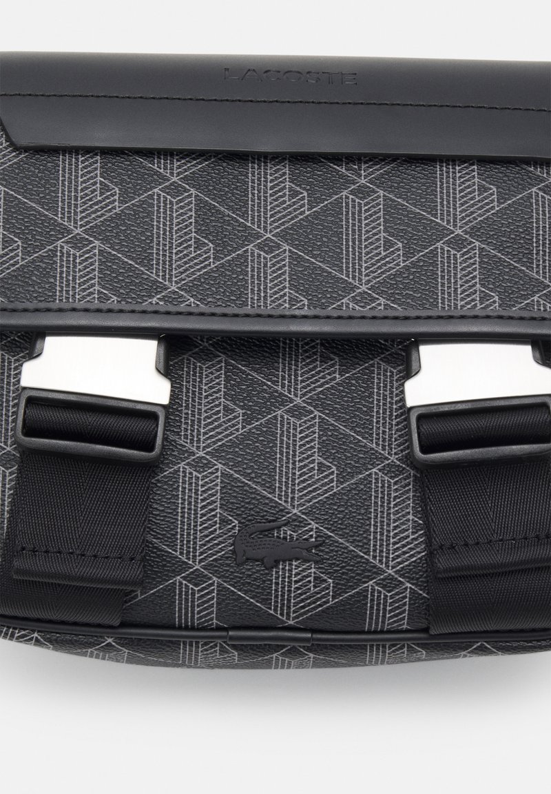 Schwarze texturierte Tasche mit geometrischem grauen Muster, ausgestattet mit silbernen Hardware-Elementen und verstellbaren Trägern. Markenlogo auf der Oberfläche geprägt.