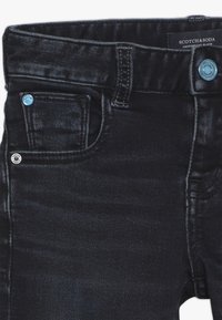 Primer plano de unos vaqueros de mezclilla azul oscuro que muestra el bolsillo frontal, el lazo del cinturón, el botón de metal y la etiqueta de la marca Scotch & Soda.