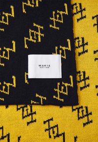 Marineblauer Strickstoff mit gelben abstrakten Mustern, versehen mit einem weißen Etikett mit "MAKIA EST. 2001 HELSINKI." Texturierter gelber Hintergrund.