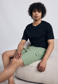 Tricou negru cu mâneci scurte asortat cu pantaloni scurți texturați, de culoare verde deschis, care au un logo, așezat pe un pouf moale, de culoare deschisă.