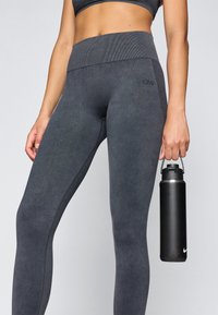 Κάπνισες γκρι κολάν με ψηλή μέση, υφή υφάσματος και πλαϊνές ραφές, συνδυασμένα με ένα μαύρο μεταλλικό μπουκάλι νερού.