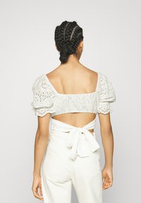 Top branco com mangas bufantes, com um design em bico e ilhós, costas expostas e detalhe de cintura amarrada, combinado com calças brancas de cintura alta.