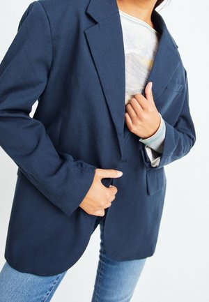 Marinblå kavaj med strukturerad design, två framfickor och enkel knappstängning. Bärs över en ljusfärgad topp och denimjeans.