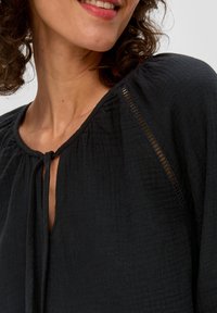 Czarny top z teksturowanej tkaniny, z dekoltem w kształcie łezki i wiązaniem. Zawiera delikatne pionowe przeszycia na ramionach.
