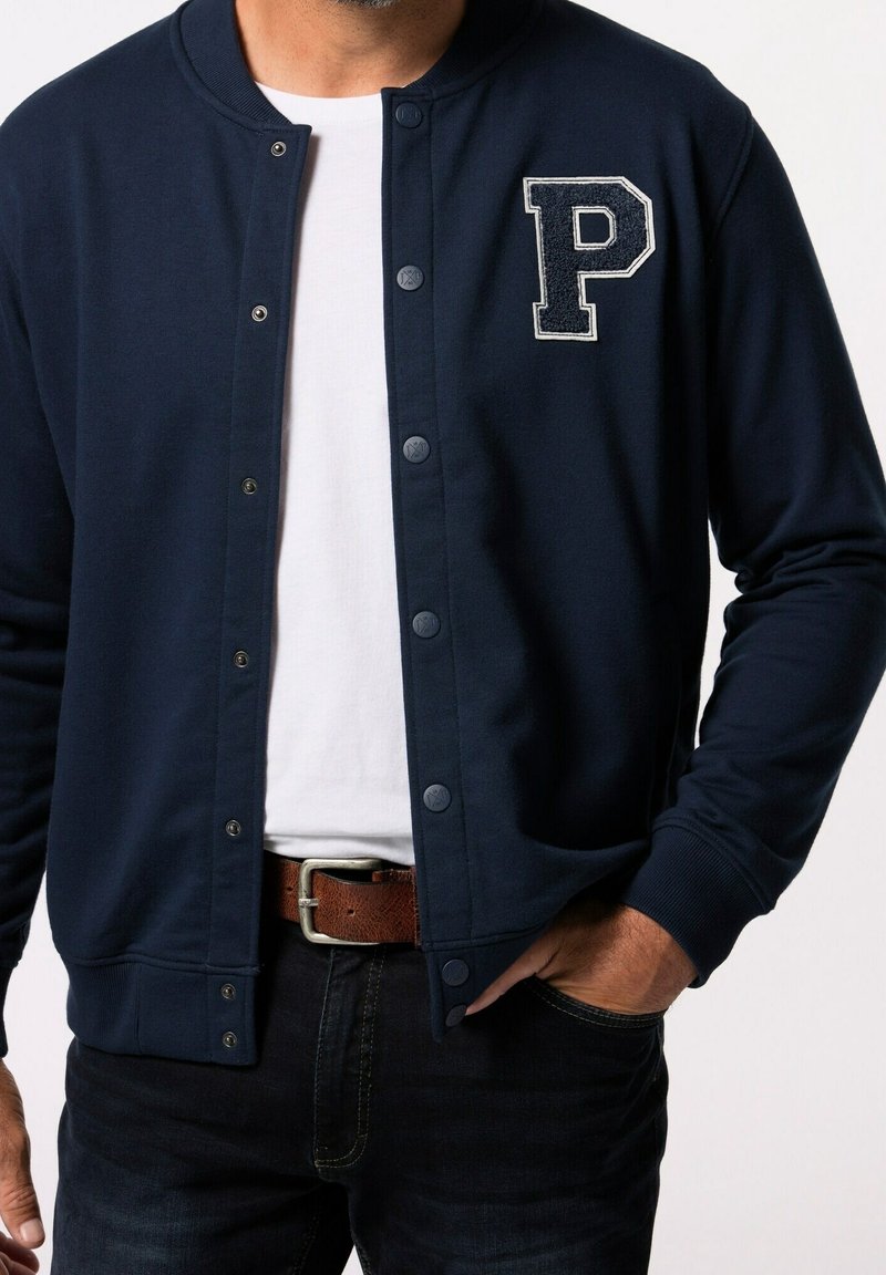 Homme portant une veste boutonnée bleu marine avec un écusson blanc en forme de "P" sur une chemise blanche, un jean foncé et une ceinture en cuir marron.