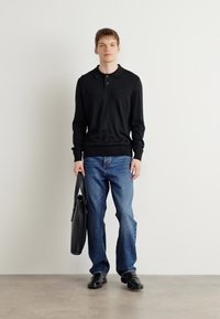 Schwarzes Langarm-Polohemd mit Kragen, kombiniert mit blauen Jeans. Hält eine schwarze Lederhandtasche und trägt schwarze Schuhe. Neutraler Hintergrund.