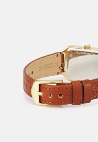 Bracelet de montre en cuir marron avec boucle en acier inoxydable doré et boîtier rectangulaire. L'arrière présente un verre transparent et des détails de marque gravés.