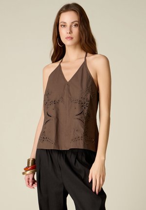 Jeune femme portant un top halter marron brodé, un pantalon noir, de grandes boucles d'oreilles anneaux et des bracelets métalliques et en bois empilés.