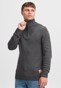 Grå strikket sweater med høj ribbet krave, der har et struktureret mønster og et lille logo-patch på nederste venstre side.