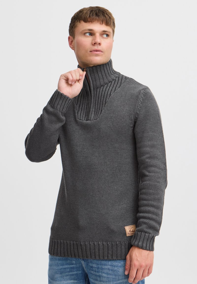 Grå strikket sweater med høj ribbet krave, der har et struktureret mønster og et lille logo-patch på nederste venstre side.