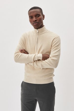Beige zip-up sweater with a soft, textured finish and fitted cuffs, paired with dark trousers. Minimalist design, suitable for casual wear.