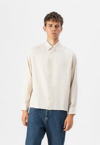 Light beige, textured long-sleeve shirt with a button-down collar, featuring front buttons and a straight hem. Paired with blue denim jeans.